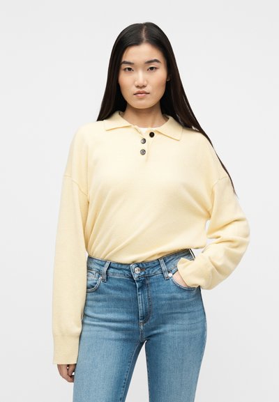 Νεαρή γυναίκα με μακριά σκούρα μαλλιά, που φοράει ανοιχτόκκινο κίτρινο πουλόβερ με τρία κουμπιά και μπλε ψηλόμεσο τζιν, με το χέρι στην τσέπη.