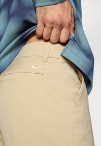 Pantalones Nike beige con una textura suave, que presentan un pequeño logotipo en el bolsillo y un diseño recto. La tela de la camisa es azul y texturizada.