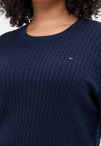 Donkerblauwe gebreide trui met een geprofileerd kabelpatroon, ronde halslijn en een klein logo in rood, wit en blauw op de borst.