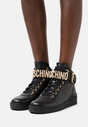 Černé kožené vysoké tenisky s zlatými očky a přezkou, s perlovým páskem, na kterém je nápis "MOSCHINO", na nohou osoby.
