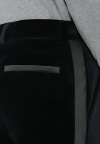 Detailné zobrazenie čiernych zamatových nohavíc so saténovo lemovanou zadnou vreckom a bočnými pruhmi, ktoré sú kombinované s svetlošedým topom.