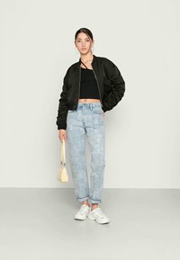 Casaco bomber preto com punhos canelados, top preto curto, jeans em patchwork azul claro, ténis brancos e uma bolsa amarela.