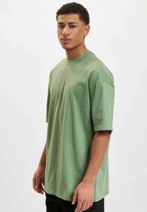 Grünes Baumwoll-T-Shirt mit kurzen Ärmeln, Rundhalsausschnitt, lässiger Passform und glatter Textur. Das Modell trägt schwarze Hosen und steht im Profil.