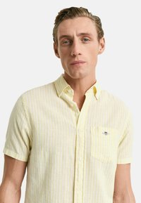 Camicia a maniche corte con bottoni di colore giallo chiaro a righe verticali bianche. Presenta una tasca frontale e un piccolo logo sul petto.
