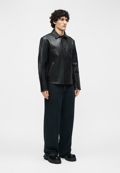 Άνδρας στέκεται φορώντας μαύρο δερμάτινο σακάκι με κουμπιά, φαρδιά σκούρα παντελόνια και μαύρα χοντρά παπούτσια μπροστά σε απλό λευκό φόντο.