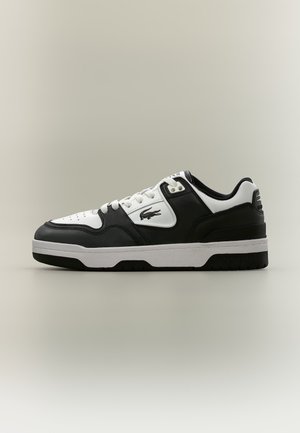 Baskets basses noir et blanc avec lacets blancs, bout perforé et logo crocodile sur le côté, présentées sur un fond uni.
