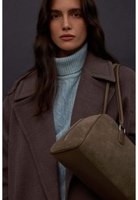 Brązowa torebka z filcu o gładkiej fakturze, zaokrąglonym dnie i skórzanymi detalami. Nosi ją w połączeniu z szarym płaszczem i niebieskim swetrem z golfem.