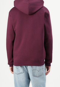 Sudadera burdeos con capucha, dobladillo y puños acanalados, confeccionada con un material suave, llevada sobre unos vaqueros azul claro con bolsillos traseros visibles.