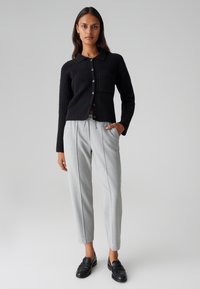 Schwarze gerippte Strickjacke mit Kragen und Fronttasche, kombiniert mit hellgrauen schmal geschnittenen Hosen. Schwarze Loafers vervollständigen das Outfit.