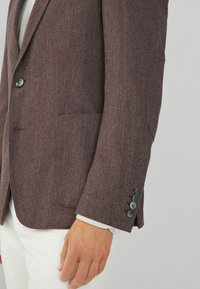 Blazer marrone a spina di pesce con una vestibilità su misura, caratterizzato da una tasca sul petto e cinque bottoni scuri sul polsino, abbinato a una camicia chiara.