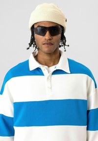 Polo à manches longues avec rayures horizontales bleues et blanches, col blanc, et patte de boutonnage à trois boutons. Porté avec un bonnet tricoté crème.