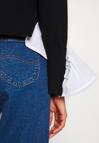 Gros plan sur une personne portant un jean bleu avec une poche arrière, un pull à manches longues noir et un poignet de chemise blanc visible sous la manche.