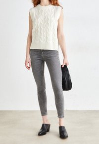 Suéter blanco sin mangas con diseño de punto de cable texturizado, combinado con jeans ajustados grises y mules negros de tacón, sosteniendo un bolso negro.