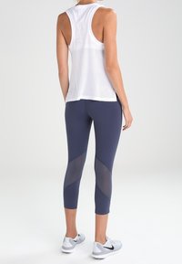 Valkoinen verkkopaitatoppi, tummansiniset leggingsit läpinäkyvillä paneeleilla ja harmaat urheilukengät. Sisältää urheilullisen selkädesignin ja istuvan siluetin.