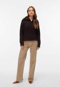 Tmavě hnědý žebrovaný pletený svetr s polovičním zipem u límce, doplněný béžovými kalhotami s širokými nohavicemi a hnědými botami na podpatku se špičatou špičkou. Jednoduchý a texturovaný design.