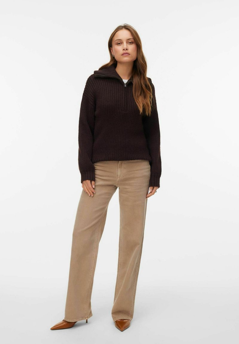 Tmavě hnědý žebrovaný pletený svetr s polovičním zipem u límce, doplněný béžovými kalhotami s širokými nohavicemi a hnědými botami na podpatku se špičatou špičkou. Jednoduchý a texturovaný design.