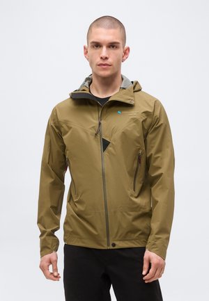 Young man with short hair wearing a brown hooded waterproof jacket with zipped pockets, standing against a plain light background.