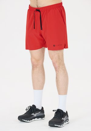Mand iført røde atletikshorts med sort snøre, hvide mellemlange sokker og sorte løbesko står mod en ensfarvet baggrund.