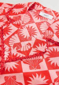 Rød og pink mønstret stof med et ternet design, der har muslingeskaller og blomstermotiver. Kanterne har farverig stikning. Mærkets label er synlig.