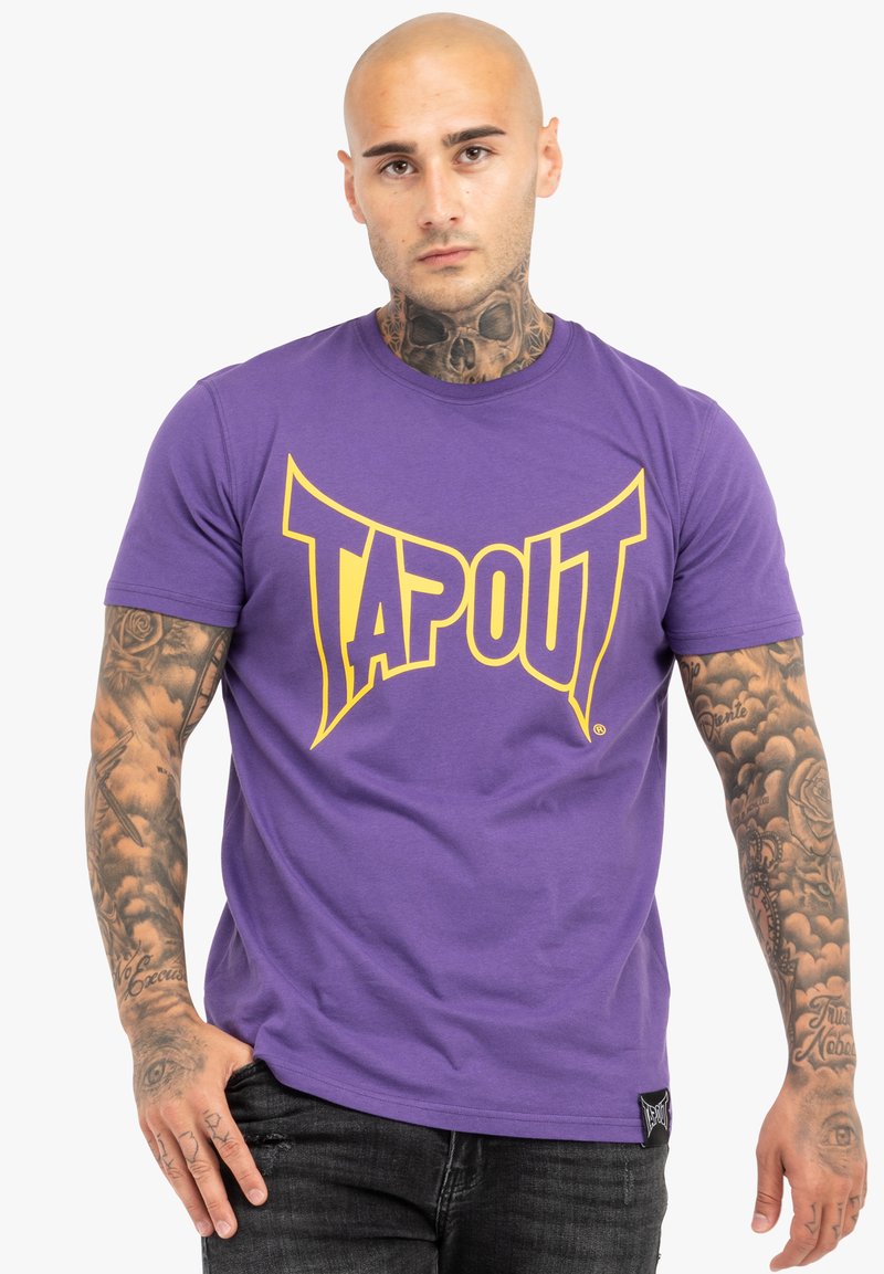 Lila pamut póló, a fronton egy nagy sárga "TAPOUT" logóval. Rövid ujjú és kerek nyakú dizájn, sötét farmernadrággal párosítva.