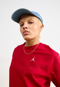 Raudona medvilninė marškinė su baltu Jumpman logotipu, derinami su džinsiniu beisbolo kepuraitė, turinčia plokščią brinklį ir siuvinėtus akcentus. Auksinė grandinėlė.