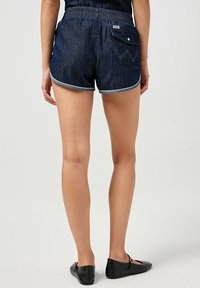 Pantaloni denim corti con elastico in vita, colore blu scuro e cuciture bianche a contrasto. La tasca posteriore presenta una chiusura a scatto e dettagli di design.
