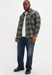 Groene en blauwe geruite flanellen blouse met twee borstzakken over een wit t-shirt, gecombineerd met blauwe jeans en zwarte laarzen.