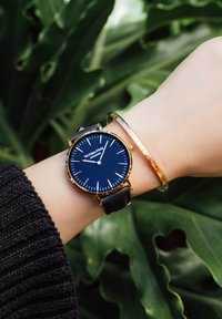 Montre avec bracelet en cuir noir, boîtier doré et cadran bleu foncé. Elle présente des index et des aiguilles blancs, accompagnés d'un bracelet doré fin.