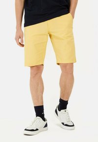 Lysgule shorts med en glat overflade, lige pasform og sidelommer, parret med hvide og marineblå sneakers og sorte sokker.