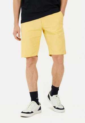 Lichtgele shorts met een gladde textuur, rechte snit en zijzakken, gecombineerd met witte en marineblauwe sneakers en zwarte sokken.