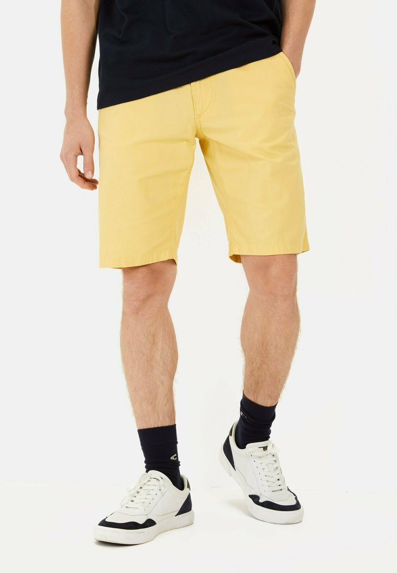 Shorts jaune clair avec une texture lisse, coupe droite et poches latérales, associés à des baskets blanches et bleu marine et des chaussettes noires.