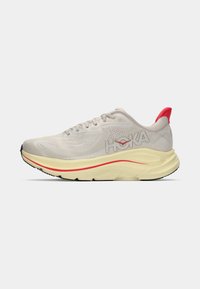Scarpa da corsa grigia chiara con tomaia in rete, logo HOKA, intersuola gialla e accentuazione rossa sul tallone. Presenta una punta arrotondata e una suola texture.