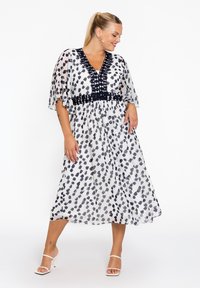 Witte midi-jurk met zwarte abstracte vlekken, doorzichtige flutter-mouwen en een zwarte versierde taille. Gestyled met witte sleehakken.