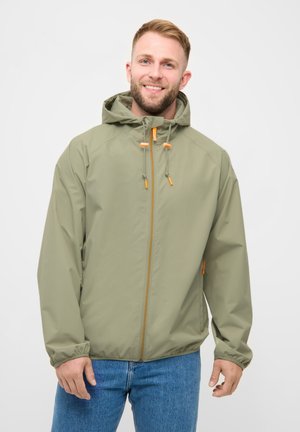 Smilende mand iført en lys olivengrøn hættetrøje med orange lynlåse kombineret med blå jeans mod en ensfarvet baggrund.