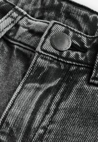 Mustmust ja hall denimkangas, millel on tekstuuriline muster, sisaldab metallnuppu, millel on graveeritud logo ja kontrastne õmblus.