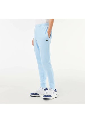 Person iført lyseblå joggingbukser med sidelommer og hvide sneakers med marineblå detaljer, står mod en ensfarvet hvid baggrund.