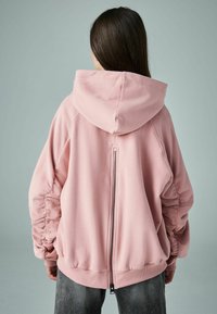 Růžová oversized mikina vyrobená z měkké tkaniny, s kapucí, staženými rukávy a centrálním zipem na zádech. Jednoduché žebrované manžety na spodním lemu.