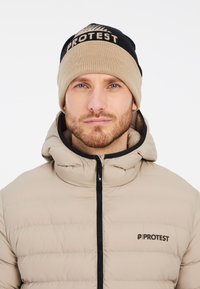 Veste en duvet beige avec des coutures horizontales et un col montant. Bonnet en tricot beige et noir avec le logo "PROTEST" sur le devant.