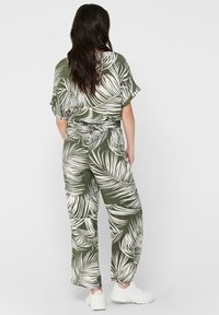 Olijfgroen jumpsuit met wit palmenbladpatroon, korte mouwen, elastische taille en wijde pijpen; gedragen met witte sneakers.
