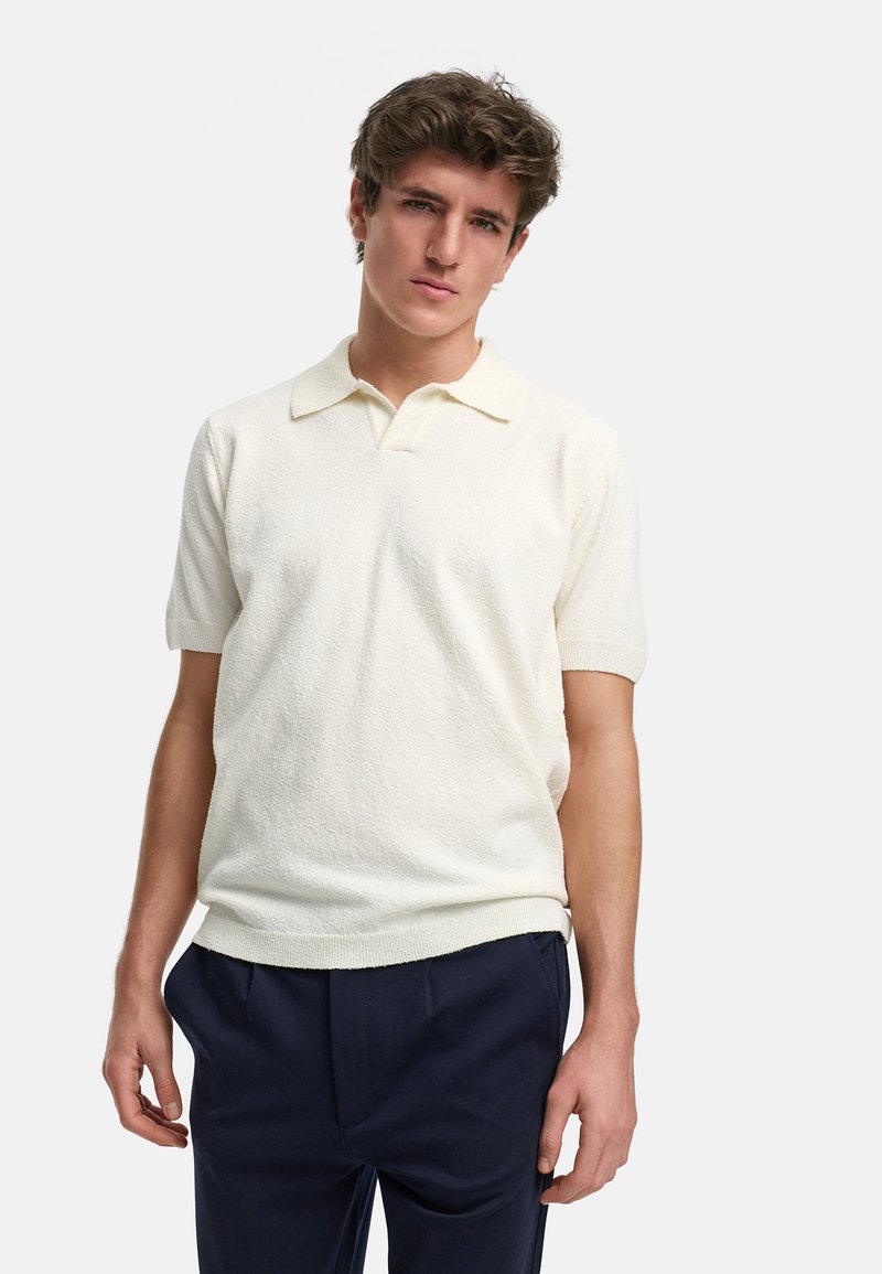 Kortermet polo skjorte i off-white strikket stoff med tekstur. Har en klassisk krage og ribbestrikkede mansjetter, sammen med marineblå bukser.