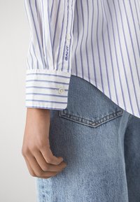 Bílá košile na knoflíky s modrými pruhy a logem GANT na rukávu, nošená s světlemodrými džínami. Má klasický límec a manžety na knoflíky.