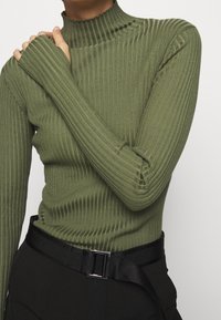 Groene geribbelde turtleneck top met lange mouwen en duimopeningen, gemaakt van rekbaar materiaal. Gedeeltelijk gedragen met een zwarte riem en hoogzittende broek.