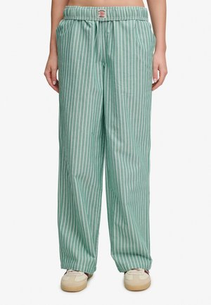 Grüne locker sitzende Hose mit weißen vertikalen Streifen, getragen mit beigen Sneakern, elastischer Bund mit kleinem vorderem Etikett.