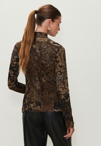 Brown long-sleeve top featuring a textured floral pattern with metallic accents. Fitted design, mock neck, and smooth fabric, paired with black pants.