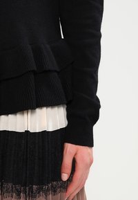 Svart stickad tröja med volangkant, bärs över en plisserad kjol i krämfärg och svart med spetsdetaljer i nederkant.