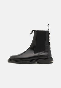 Musta nahkainen Chelsea-boot, jossa on joustavat paneelit, pyöristetty kärki, koristeellista metalliosaa takana ja teksturoitu pohja.