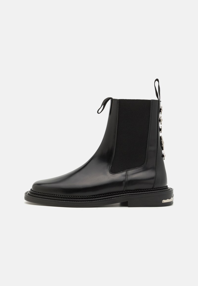 Musta nahkainen Chelsea-boot, jossa on joustavat paneelit, pyöristetty kärki, koristeellista metalliosaa takana ja teksturoitu pohja.