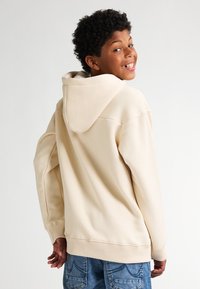 Felpa beige con cappuccio regolabile, polsini e orlo a coste; indossata con jeans in denim blu. Il tessuto appare morbido e tessurizzato.