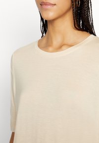 Beżowa koszulka z okrągłym dekoltem, wykonana z miękkiego, lekkiego materiału. Krótkie rękawy z luźnym krojem i delikatną teksturą. Minimalistyczny design bez wzorów.