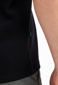 Schwarzes Baumwoll-T-Shirt mit abgerundetem Saum, versehen mit einem kleinen grünen Etikett an der Seite. Glatte Textur und kurze Ärmel.